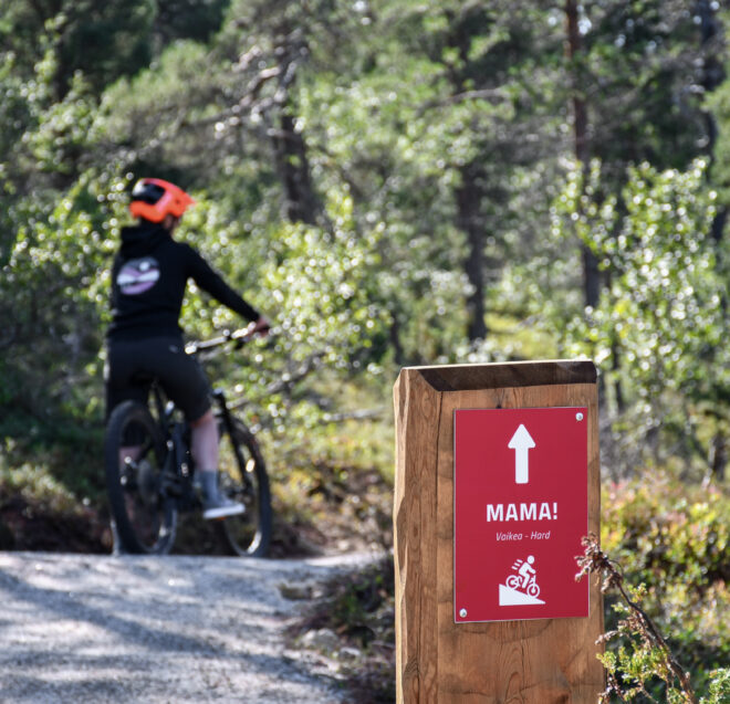 Oranssia kypärää päässään pitävä pyöräilijä ajaa metsäpolkua pitkin. Etualalla on punainen kyltti, jossa on polkupyöräkuvake ja nuoli sekä teksti ”MAMA! Ylämäki – vaikea”. Taustalla näkyy vihreitä puita ja auringonvaloa.