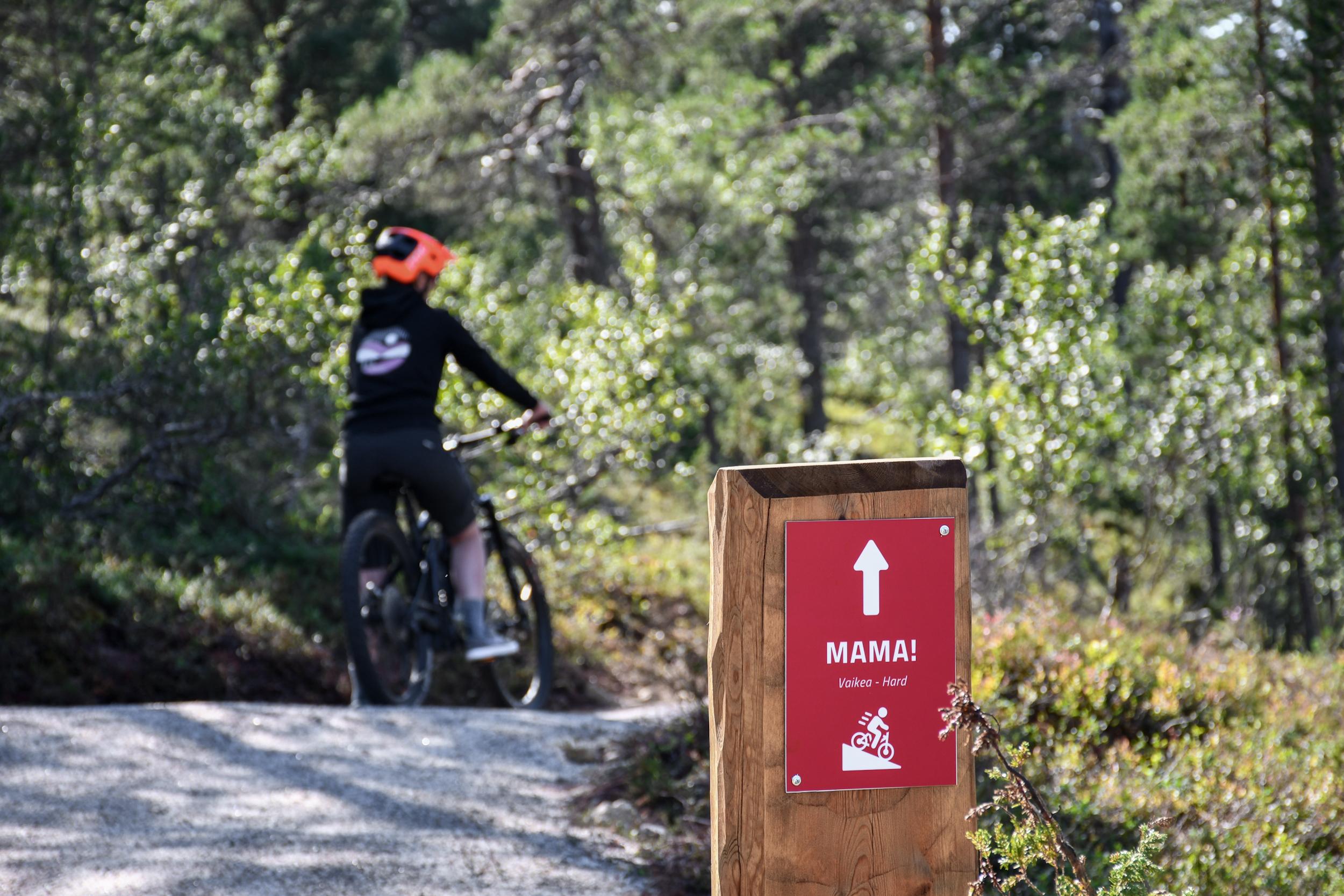 Oranssia kypärää päässään pitävä henkilö ajaa polkupyörällä metsäpolulla. Etualalla on punainen kyltti, jossa on eteenpäin osoittava nuoli ja teksti ”MAMA!”, ja taustalla näkyy vihreitä puita.