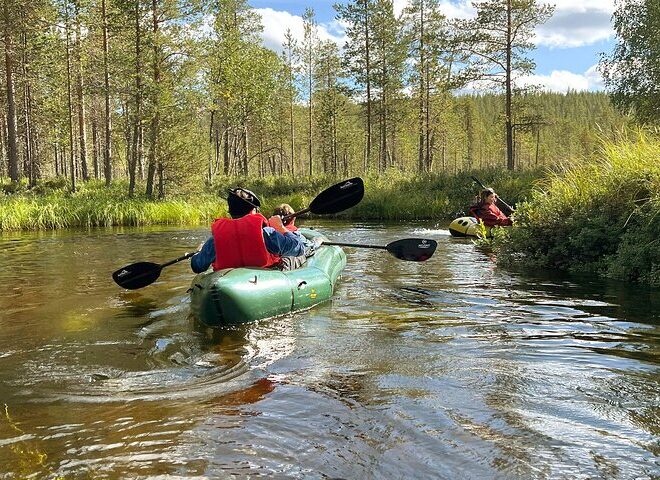 Kolme ihmistä meloo kajakilla rauhallisella joella, jota ympäröivät vihreät puut ja ruoho sinisen taivaan alla, jossa on hajanaisia pilviä. Kaksi heistä on vihreässä kajakissa, yksi keltainen kajakissa, ja kaikilla on pelastusliivit päällä.