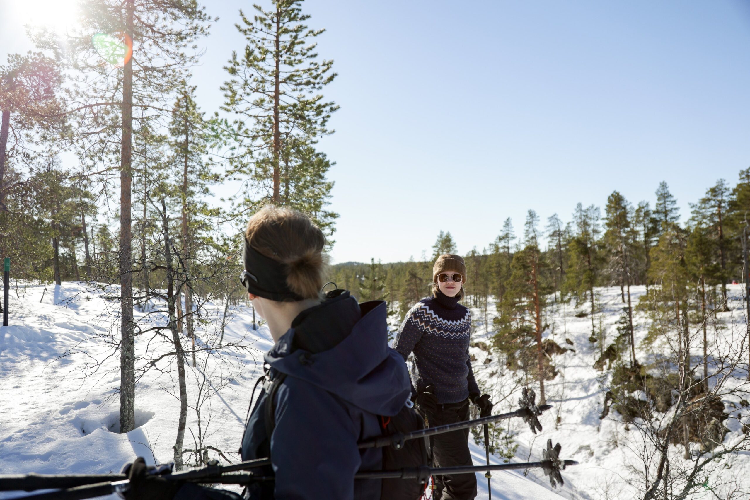 Kaksi talvivaatteisiin pukeutunutta henkilöä lumikenkäilee lumisessa metsässä aurinkoisena päivänä, puiden ja sinisen taivaan ympäröimänä.