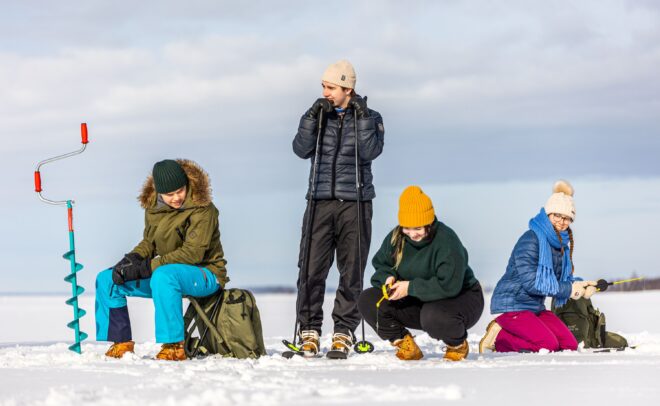 Neljä talvivaatteisiin pukeutunutta henkilöä istuu ja polvistuu lumella valmistautuen jääkalastukseen. Yksi henkilö poraa reikää porakoneella, kun taas muut käsittelevät kalastusvälineitä. Taustalla näkyy laaja luminen maisema pilvisen taivaan alla.