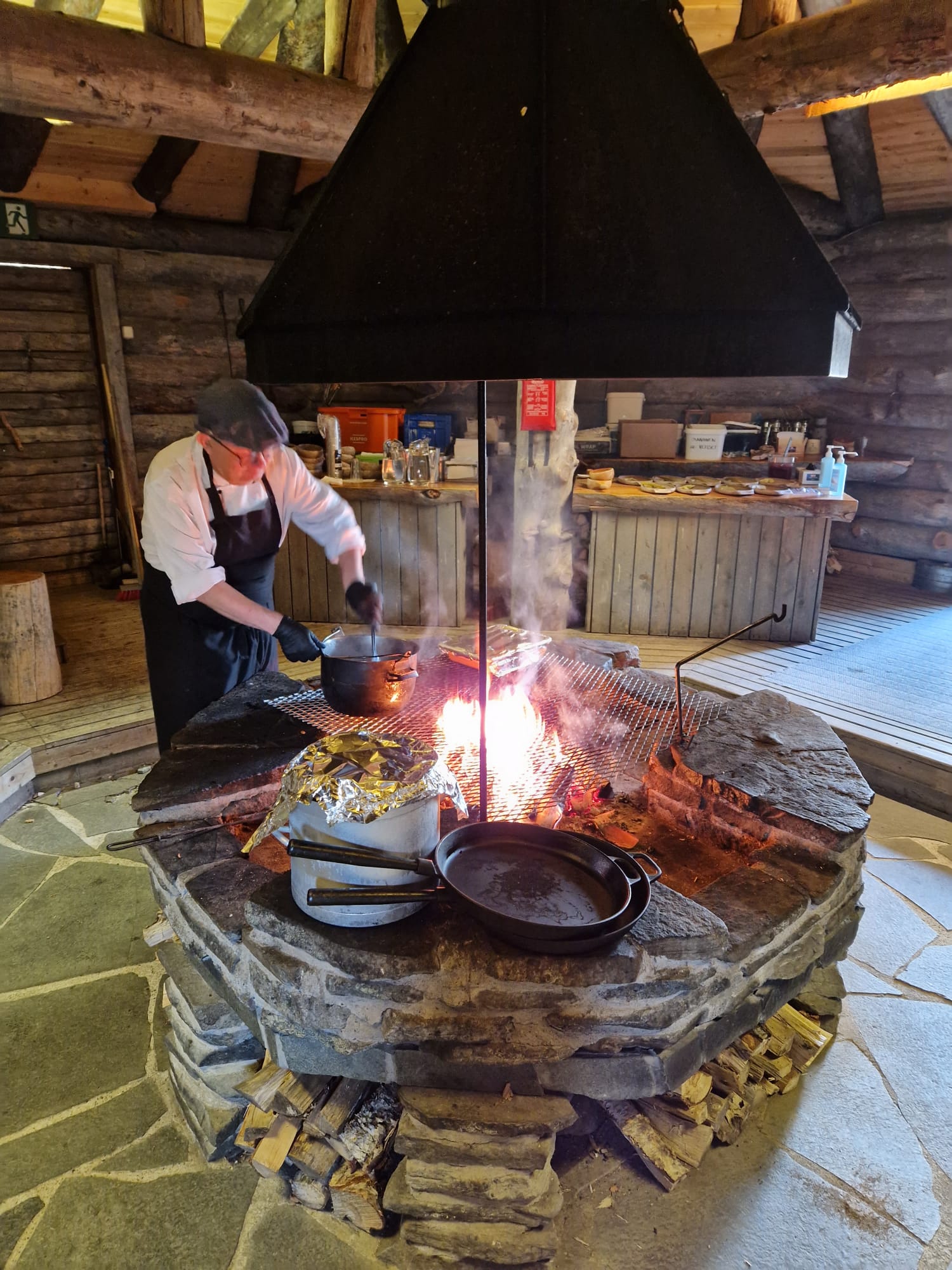 Kokki valmistaa ruokaa avotakan äärellä rustiikkisessa puumökissä. Kattilat, pannut ja ruoka paistuvat tulen päällä, ja taustalla näkyy keittiötarvikkeita ja hyllyjä.