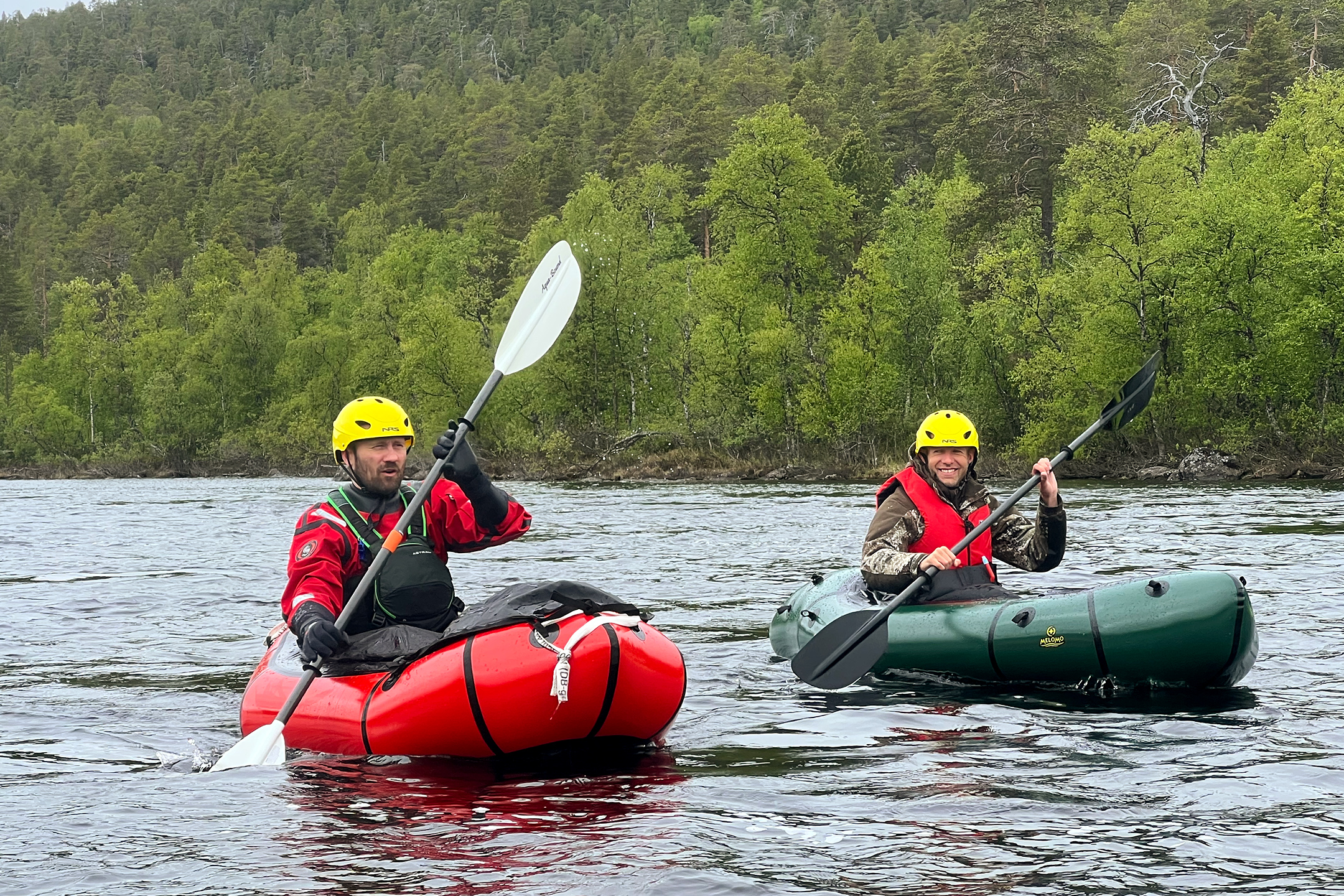 Kaksi kypärää ja pelastusliivejä käyttävää henkilöä meloo ilmatäytteisillä kajakkeilla rauhallisella joella, jota ympäröivät tiheä vihreä metsä ja kukkulat, pilvisen taivaan alla.