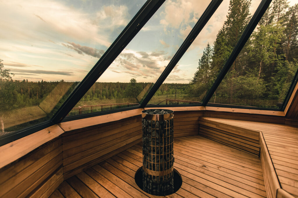 Puinen sauna, jossa on lasiseinät ja -katto sekä keskellä sijaitseva metallinen kiuas, ja josta avautuu näkymä vihreään metsään osittain pilvisen taivaan alla auringonlaskun aikaan – moderni saunapaikka, joka on suunniteltu rentoutumiseen luonnon keskellä.