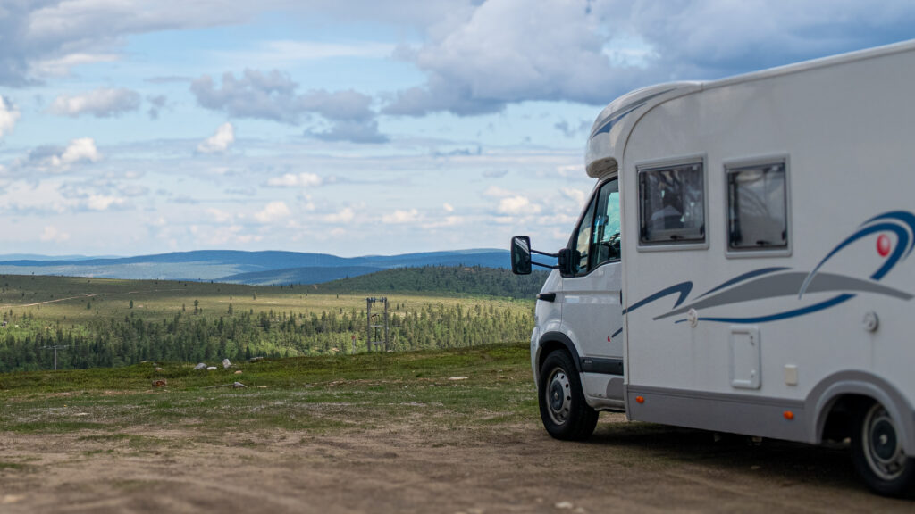 A white camper van parked on a grassy field overlooks distant green hills and forests under a partly cloudy sky, capturing the serene beauty of camping in Lapland.