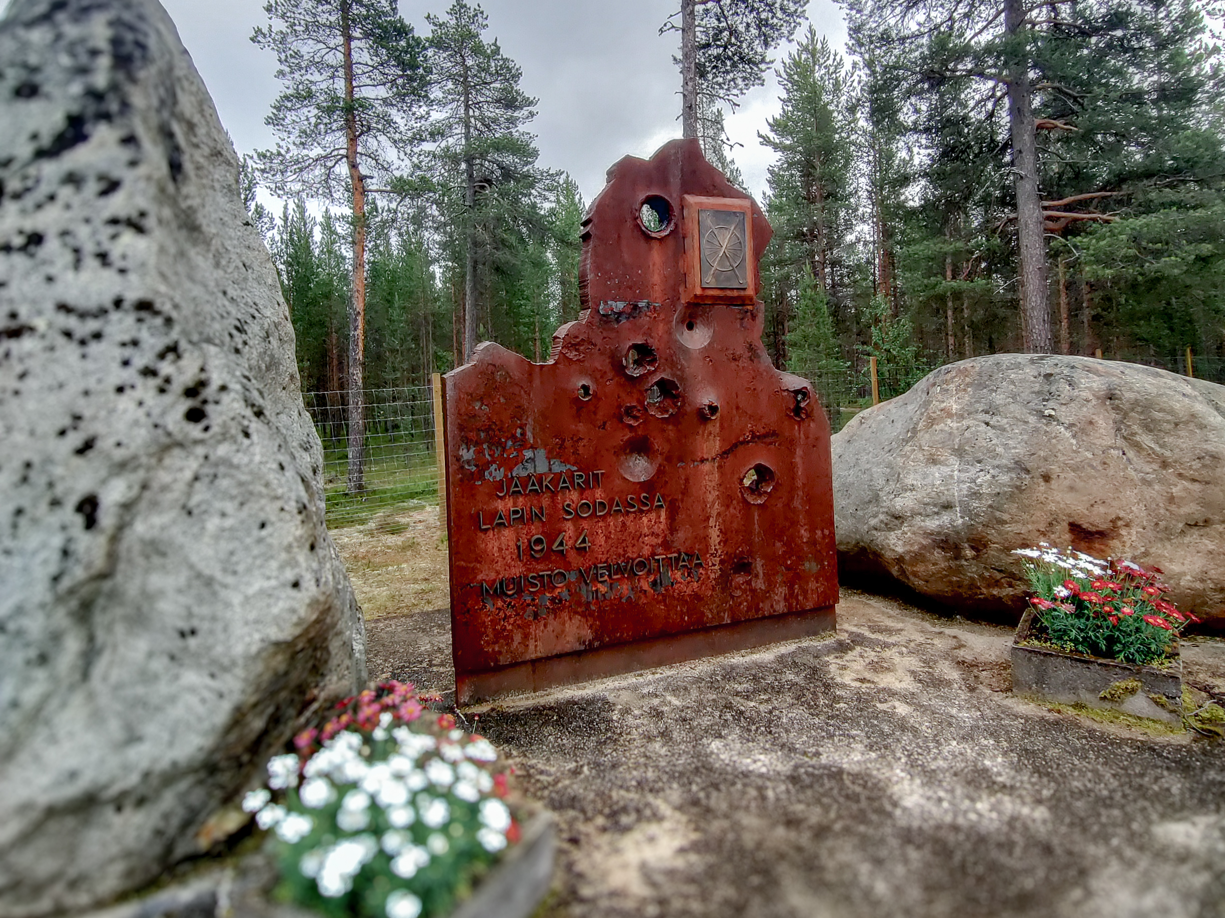 Une plaque commémorative rouillée et criblée de balles se dresse entre deux gros rochers près de Karigasniemi et Kaamanen, entourée de pins et de petits pots contenant des fleurs blanches et rouges.