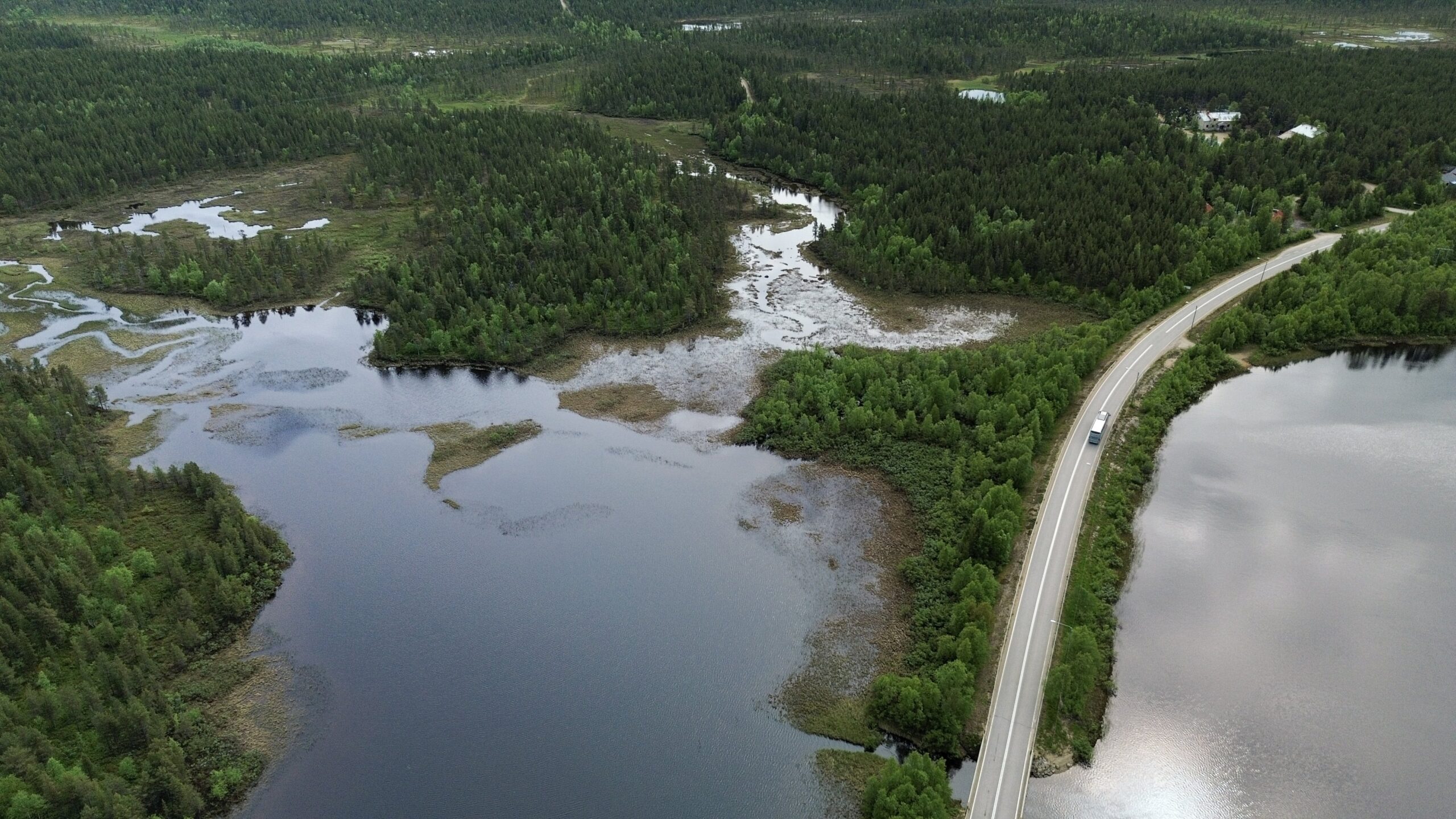 Ilmakuva mutkittelevasta tiestä rauhallisen järven rannalla, jota ympäröi tiheä vihreä metsä ja kosteikkoalueita pilvisen taivaan alla; kuvassa näkyy Karigasniemen ja Kaamanen välisen alueen rauhallinen maisema.