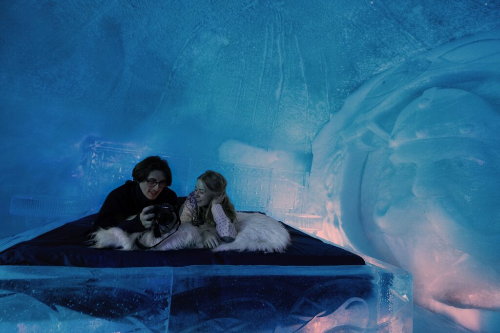 Two people relax on a bed made of ice, covered with furs, inside an ice room with blue lighting and carved ice walls, enjoying the unique experience of premium accommodation in Lapland.