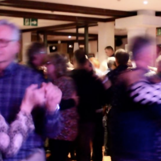 A group of people, mostly older adults, dance closely in pairs in a warmly lit indoor space. The motion makes some figures appear blurred.