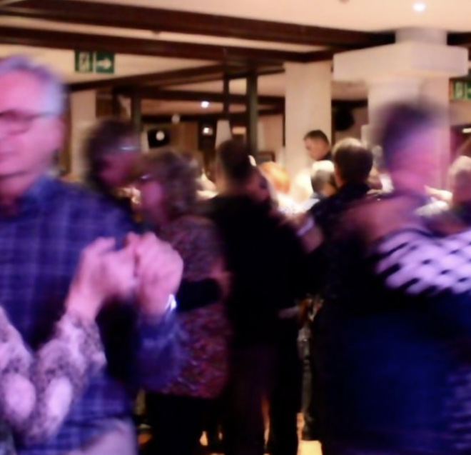 A group of people, mostly older adults, dance closely in pairs in a warmly lit indoor space. The motion makes some figures appear blurred.