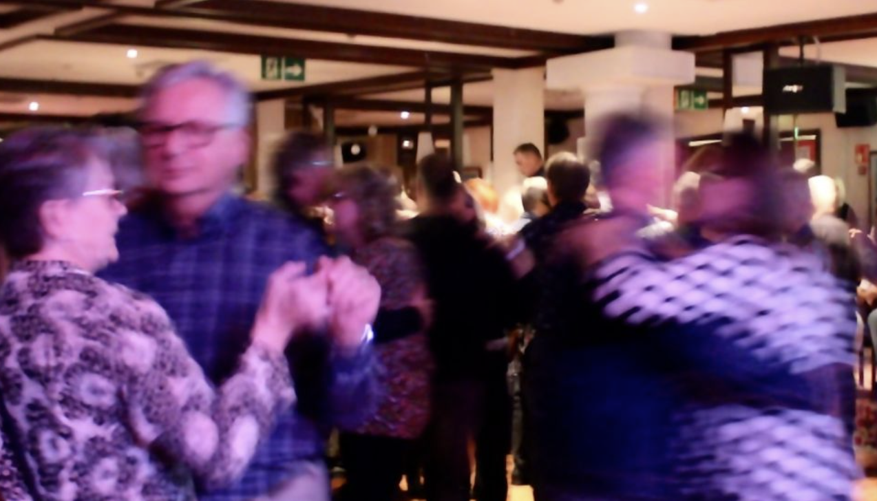 A group of people, mostly older adults, dance closely in pairs in a warmly lit indoor space. The motion makes some figures appear blurred.