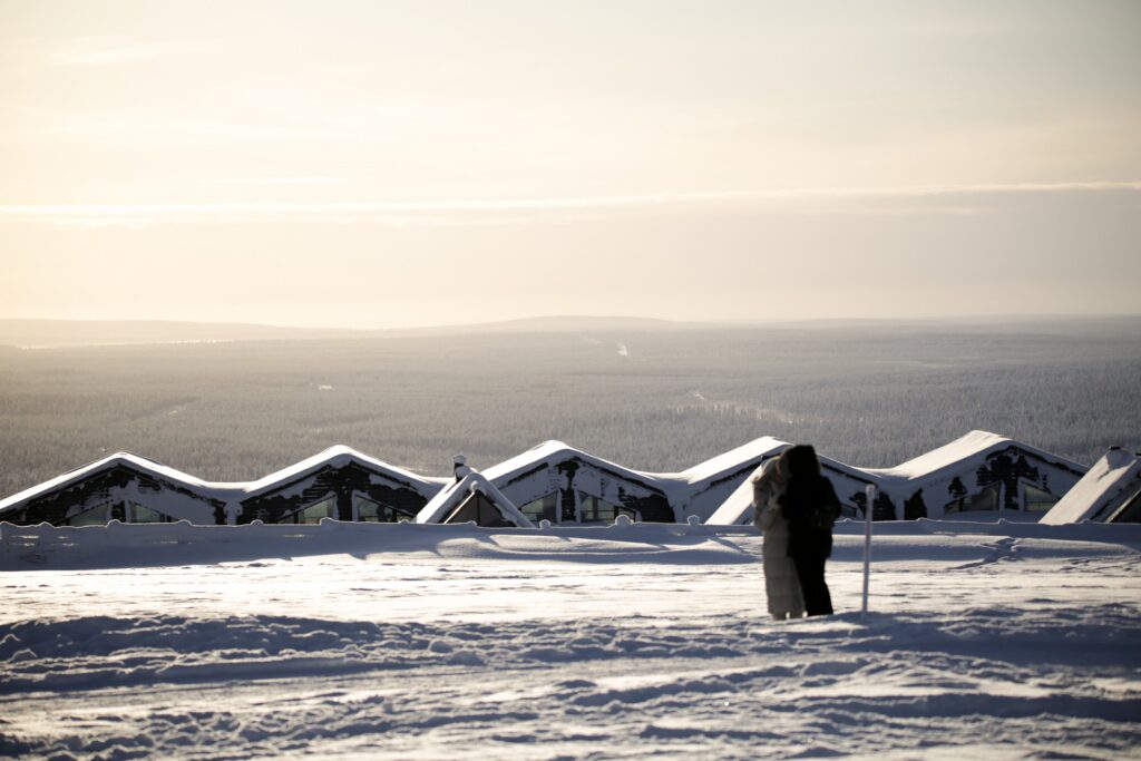 A snowy landscape with a row of snow-covered rooftops in the background—perhaps belonging to a cozy hotel in Lapland. Two people stand closely together in the foreground, looking toward the horizon under a pale sky.