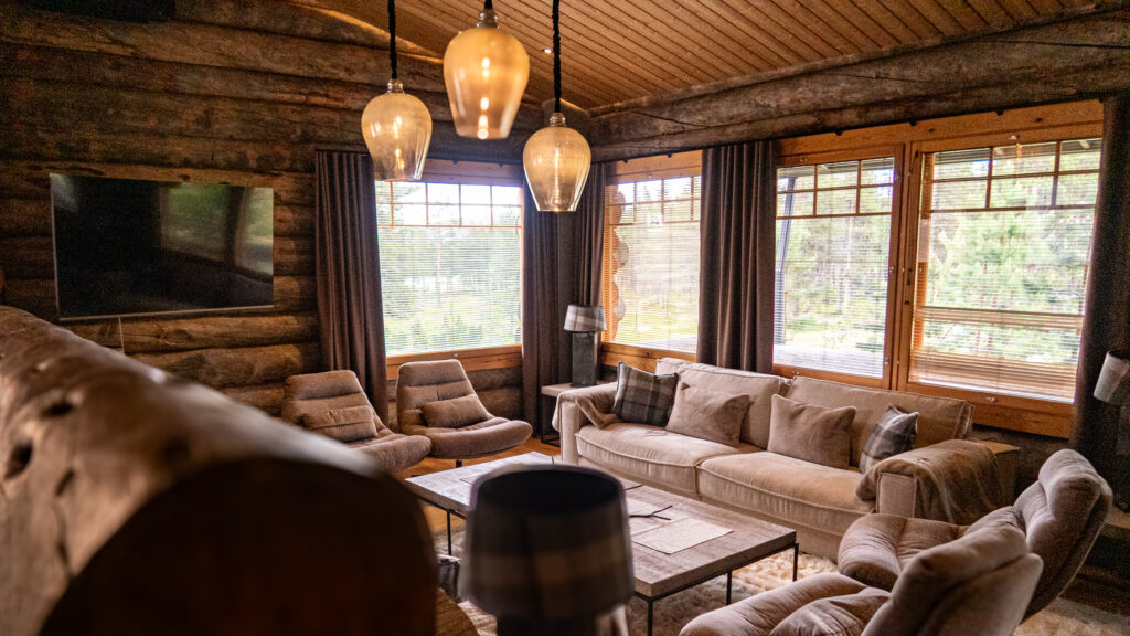 Cozy log cabin living room with large windows, beige sofas, armchairs, a wooden ceiling, hanging light fixtures, and a wall-mounted TV, overlooking green trees outside—perfect for experiencing the tranquil charm of a cabin in Lapland.