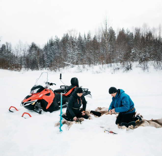 Kaksi ihmistä istuu eläinten nahkojen päällä lumessa punaisen moottorikelkan vieressä ja valmistelee ruokaa tai varusteita puiden edessä. Maisema on talvinen ja syrjäinen, ja kuvan kulmassa näkyy vihreä CO2-kompensaatiologo.