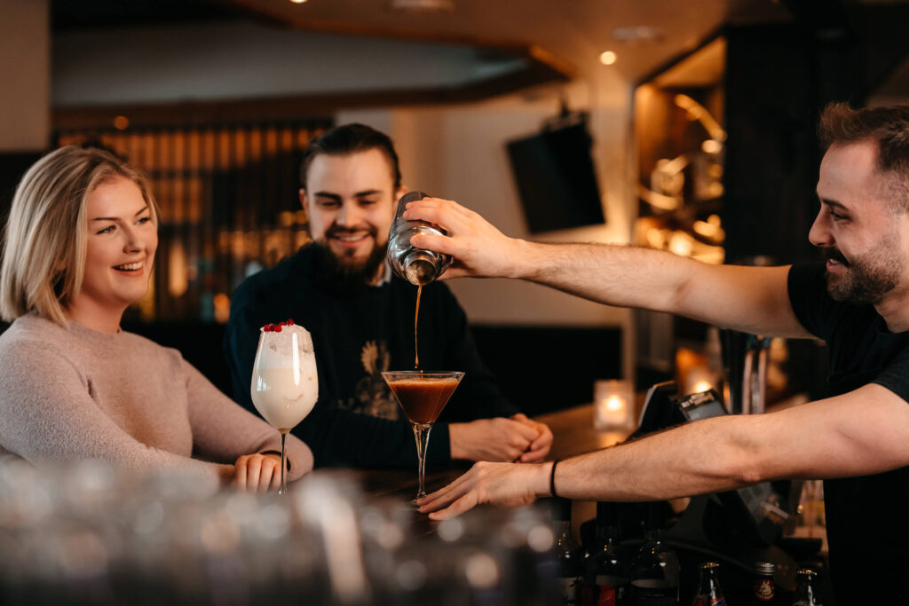 Baarimikko kaataa cocktailin martinilasiin kahdelle hymyilevälle asiakkaalle, jotka istuvat hämärästi valaistussa baarissa; toisella heistä on kädessään kermanvalkoinen juoma, jonka päällä on kirsikka.