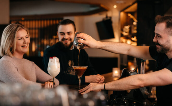 Baarimikko kaataa cocktailin martinilasiin kahdelle hymyilevälle asiakkaalle, jotka istuvat hämärästi valaistussa baarissa; toisella heistä on kädessään kermanvalkoinen juoma, jonka päällä on kirsikka.