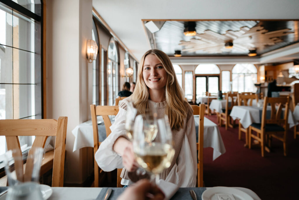 Nainen hymyilee kohottaessaan maljan valkoviiniä kädessään pöydän ääressä valoisassa, tyylikkäässä ravintolassa, jossa on puiset tuolit ja valkoiset pöytäliinat.