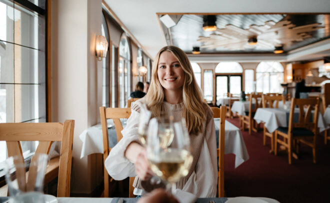 Nainen hymyilee kohottaessaan maljan valkoviiniä kädessään pöydän ääressä valoisassa, tyylikkäässä ravintolassa, jossa on puiset tuolit ja valkoiset pöytäliinat.