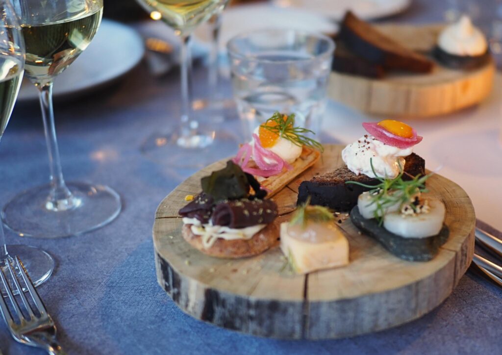 Puinen tarjoilulautanen, jolla on valikoima gourmet-alkupaloja, kuten mereneläviä, juustoa, yrttejä ja marinoituja sipuleita, ja jonka ympärillä on viinilaseja, lautasia ja ruokailuvälineitä sinisellä pöytäliinalla.