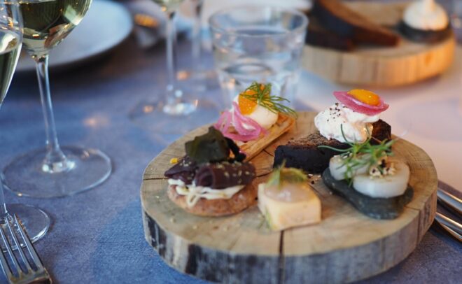 Puinen tarjoilulautanen, jolla on valikoima gourmet-alkupaloja, kuten mereneläviä, juustoa, yrttejä ja marinoituja sipuleita, ja jonka ympärillä on viinilaseja, lautasia ja ruokailuvälineitä sinisellä pöytäliinalla.