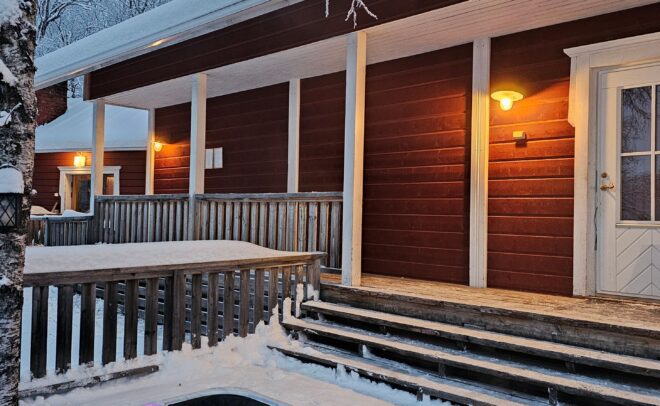 A snow-covered wooden house with warm lights, front steps, and a hot tub glowing with purple lights in the snowy yard, surrounded by trees in winter.