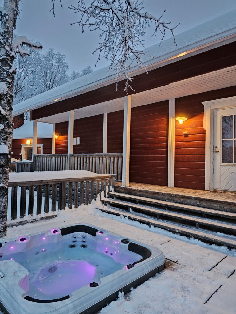 A snow-covered wooden house with warm lights, front steps, and a hot tub glowing with purple lights in the snowy yard, surrounded by trees in winter.