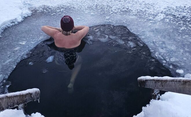 A person wearing a red hat relaxes in an ice hole surrounded by snow, with arms resting on the edge of the ice and wooden steps leading down to the frozen water.