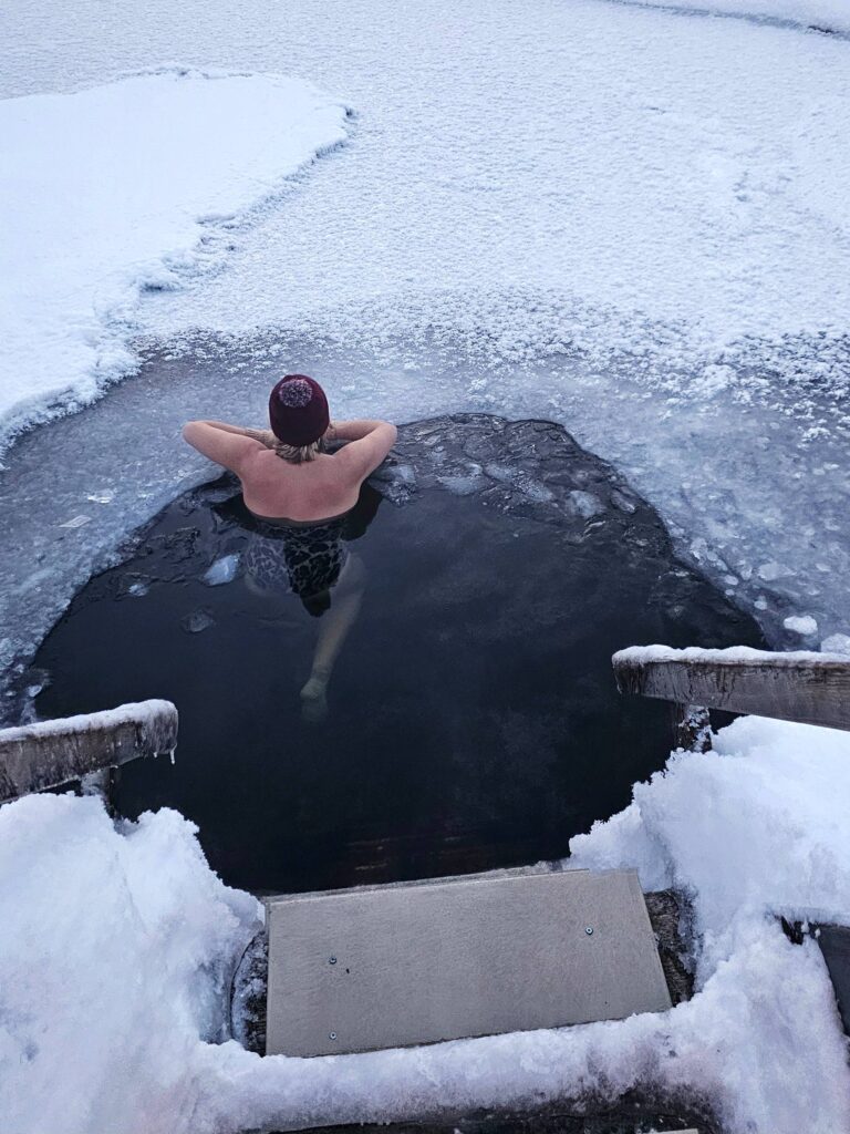 A person wearing a red hat relaxes in an ice hole surrounded by snow, with arms resting on the edge of the ice and wooden steps leading down to the frozen water.