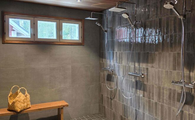Modern shower room with tiled walls and floor, dual showerheads, a wooden bench with a woven basket, small windows, and a wood-paneled ceiling with recessed lights.
