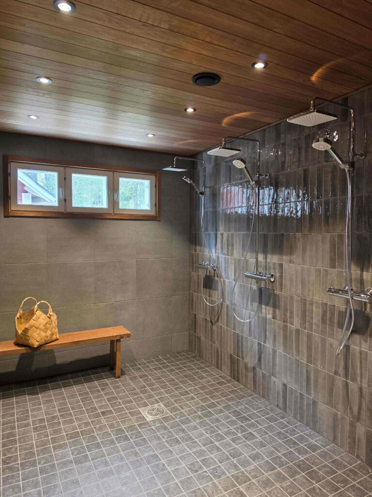 Modern shower room with tiled walls and floor, dual showerheads, a wooden bench with a woven basket, small windows, and a wood-paneled ceiling with recessed lights.