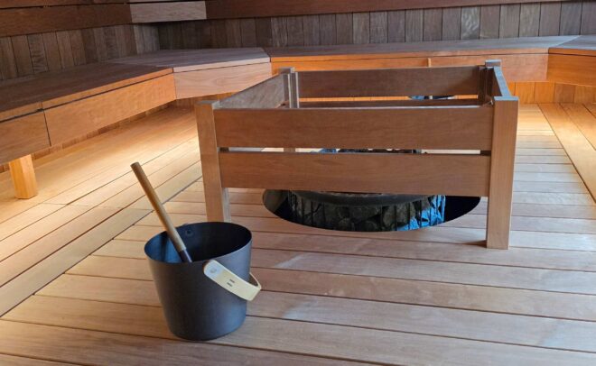 A modern wooden sauna room with benches along the walls, a central heater surrounded by a wooden frame, and a black bucket with a wooden ladle placed on the floor in the foreground.
