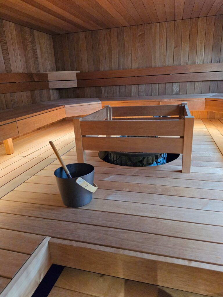 A modern wooden sauna room with benches along the walls, a central heater surrounded by a wooden frame, and a black bucket with a wooden ladle placed on the floor in the foreground.