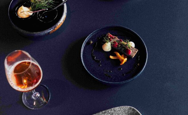 Elegant dishes with colorful vegetables and garnishes are arranged on dark plates along with a glass of rosé wine, all set against a deep blue table background.