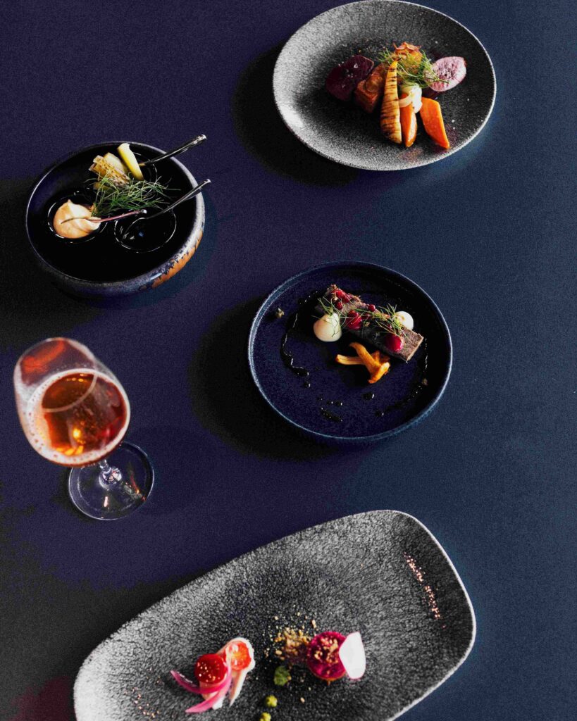 Elegant dishes with colorful vegetables and garnishes are arranged on dark plates along with a glass of rosé wine, all set against a deep blue table background.