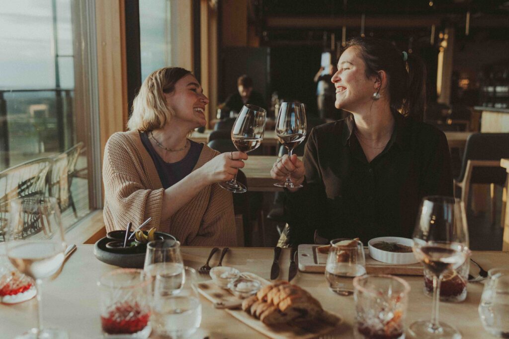 Two women sit at a restaurant table, smiling and clinking wine glasses together. The table is set with food, drinks, and bread, with sunlight streaming in through large windows behind them.