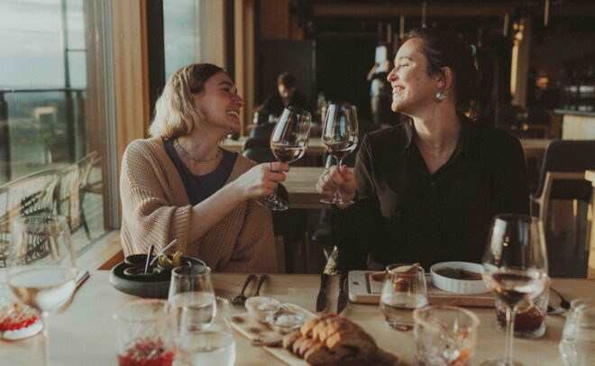 Two women sit at a restaurant table, smiling and clinking wine glasses together. The table is set with food, drinks, and bread, with sunlight streaming in through large windows behind them.