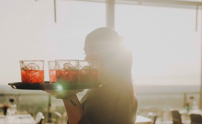 A server carries a tray with red drinks in glasses inside a sunlit restaurant, with sunlight streaming through large windows in the background.