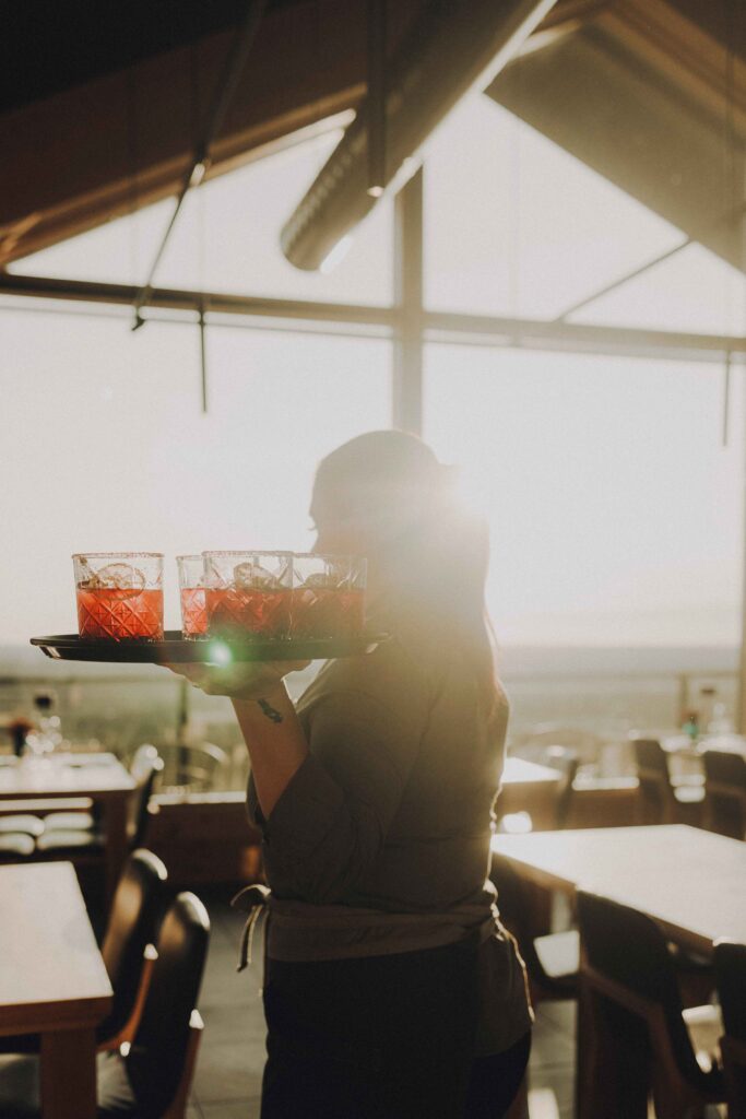 A server carries a tray with red drinks in glasses inside a sunlit restaurant, with sunlight streaming through large windows in the background.