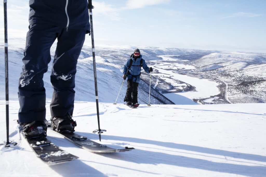 Two people skiing on a snowy mountain in Utsjoki Lapland, with distant snow-covered hills and the winding River Teno visible below. The closer person’s skis and poles are clearly seen in the foreground.
