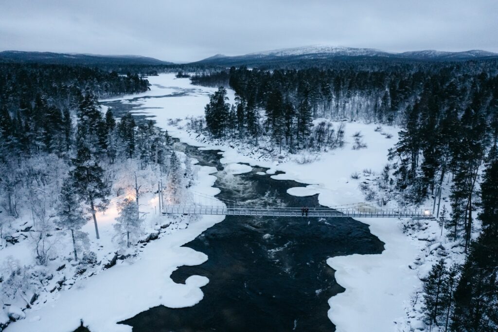 Luftaufnahme eines verschneiten Inari-Waldes, durch den ein Fluss fließt. Eine Hängebrücke überspannt den Fluss, umgeben von schneebedeckten Bäumen und eisigem Wasser, im Hintergrund sind Berge unter einem bewölkten Himmel zu sehen.