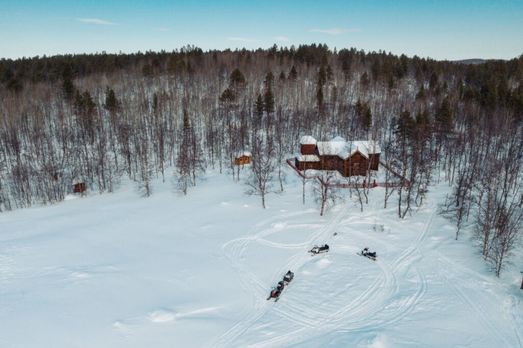 Eine Blockhütte, umgeben von einem verschneiten Wald in Inari, mit Schneemobilen und Schlitten auf Spuren im Schnee, unter einem strahlend blauen Himmel.
