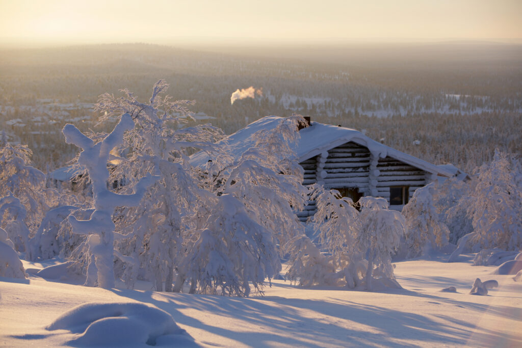 Luminen hirsimökki, jonka savupiipusta nousee savua, sijaitsee paksun lumipeitteen alla olevien puiden keskellä, peitettynä pehmeään kultaiseen auringonvaloon talvimaisemassa, joka muistuttaa Lapin mökkejä.