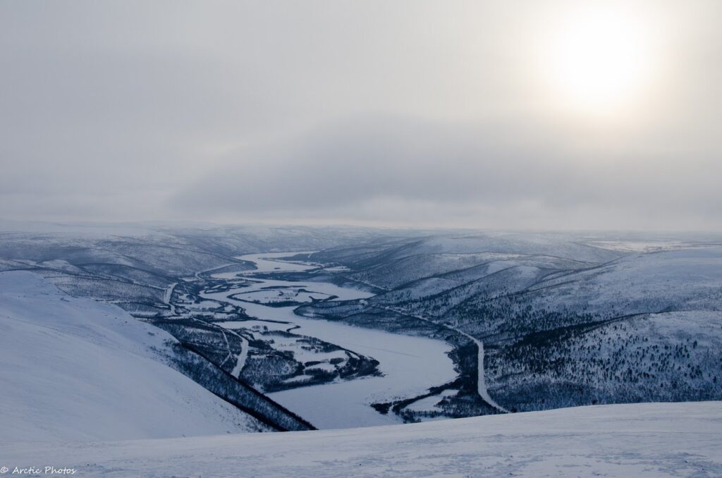 A winding River Teno flows through snow-covered hills under a cloudy, overcast sky with the sun faintly visible, creating a cold, serene winter landscape in Utsjoki Lapland.