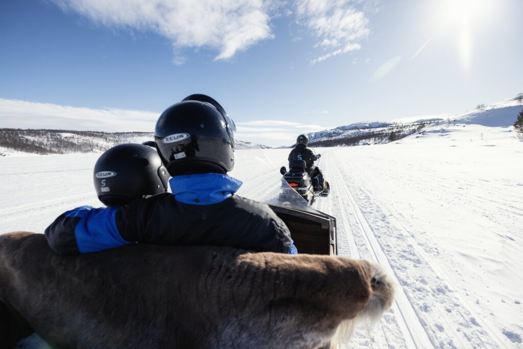 Zwei Menschen mit Helmen sitzen in einem Schlitten, einer hat seinen Arm um den anderen gelegt; der Schlitten wird von einem Schneemobil durch eine verschneite Landschaft unter strahlend blauem Himmel in der malerischen Region Kirkenes gezogen. Der Schlittensitz ist mit einem Rentierfell bedeckt.