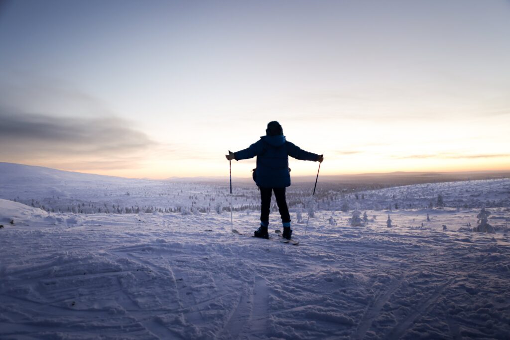 Eine Person in Winterkleidung steht mit Skistöcken auf verschneitem Boden und blickt auf einen malerischen Sonnenuntergang über der weiten, schneebedeckten Landschaft von Kiilopää.