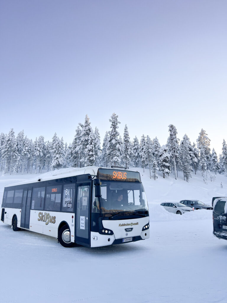 Valkoinen hiihtobussi on pysäköity lumiseen pysäköintialueeseen, jota ympäröivät lumipeitteiset puut ja useita autoja kirkkaan, vaaleansinisen taivaan alla – kuvankaunis näkymä, joka vangitsee talven taian ja antaa vinkin siitä, miten pääset Pohjois-Lappiin seuraavaa seikkailuasi varten.