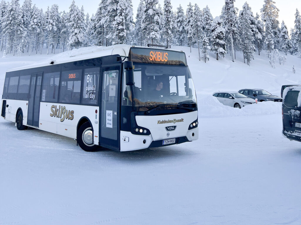 Valko-musta hiihtobussi on pysäköity talvipäivänä lumiselle pysäköintialueelle, jota ympäröivät lumipeitteiset puut ja useita pysäköityjä autoja.