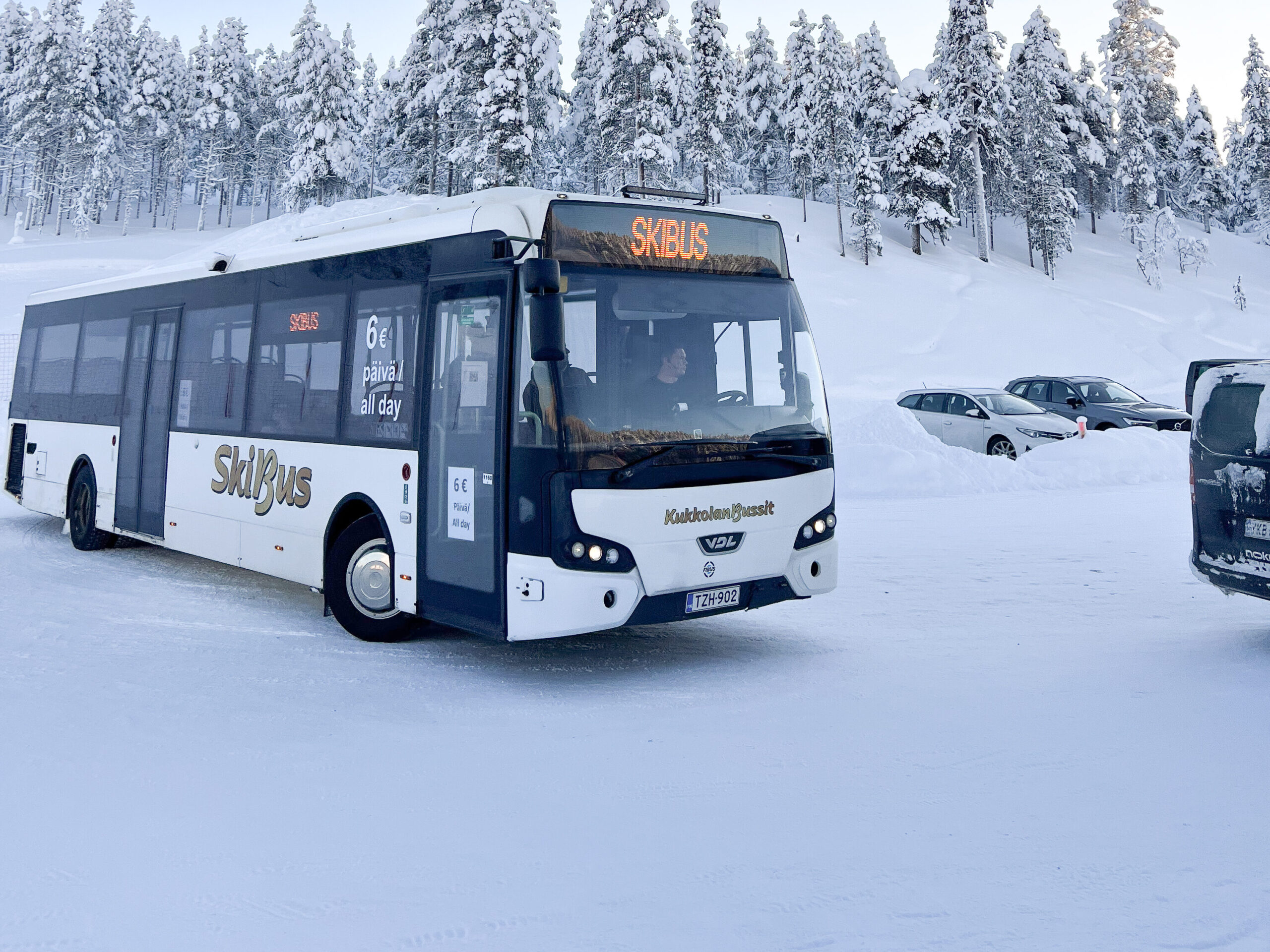 Valkoinen hiihtobussi on pysäköity lumiseen pysäköintialueeseen, jota ympäröivät lumipeitteiset puut ja useita autoja kirkkaan, vaaleansinisen taivaan alla – kuvankaunis näkymä, joka vangitsee talven taian ja antaa vinkin siitä, miten pääset Pohjois-Lappiin seuraavaa seikkailuasi varten.