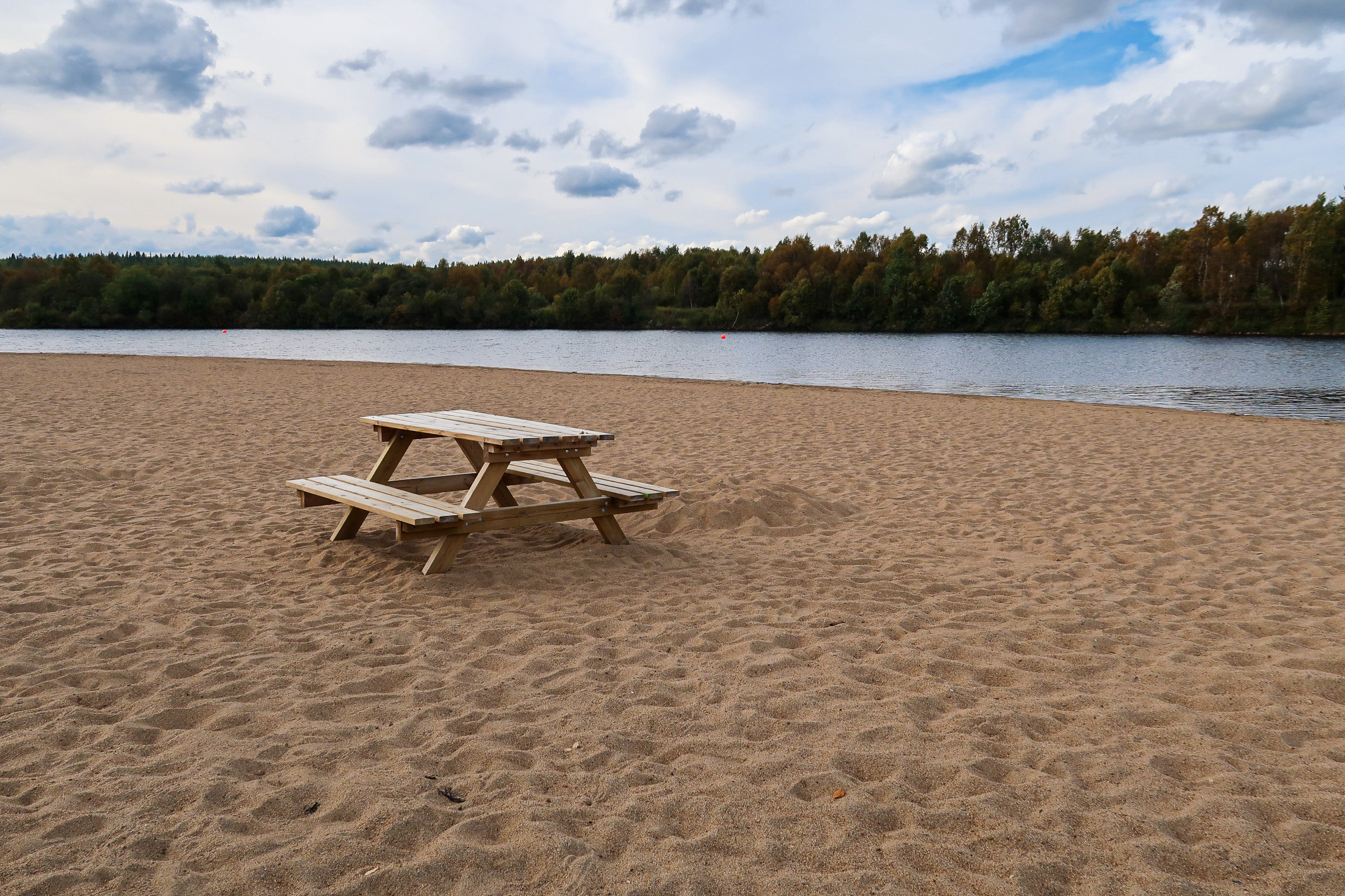 A wooden picnic table sits on a sandy beach by a calm lake near Ivalo Lapland, with trees and a cloudy sky in the background.