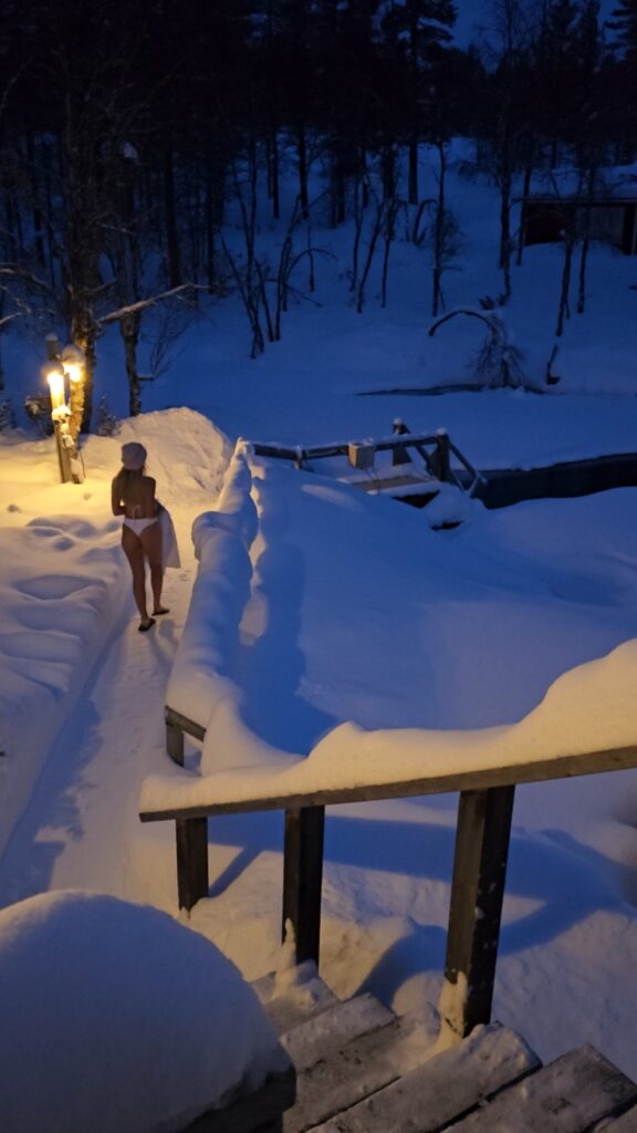 A person in a towel and hat walks outside at dusk in a snowy landscape, heading toward a wooden railing and a snow-covered area illuminated by a lamppost.